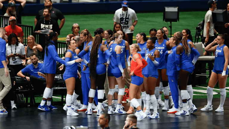 A volleyball team in blue uniforms huddles together, celebrating on the court.